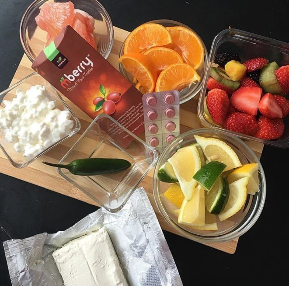 There are different types of fruits such as pink grapefruit, orange that are cut into slices, strawberries cut in half, lemon and lime cut in slices. There is also green chili, and cottage cheese in the picture frame. All the fruits, chili, and cottage cheese are put into a separate small glass bowl. They are all placed on top of the cutting board with mberry package and the tablets that display in the middle of the fruits and foods.